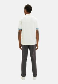Weißes Poloshirt mit kurzen Ärmeln, das blaue und hellblaue Streifenakzente an den Ärmeln aufweist, kombiniert mit dunklen Hosen und weißen Turnschuhen.