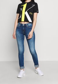 Camiseta negra corta con un gran diseño gráfico amarillo y blanco, combinada con vaqueros azules ajustados y zapatillas blancas.