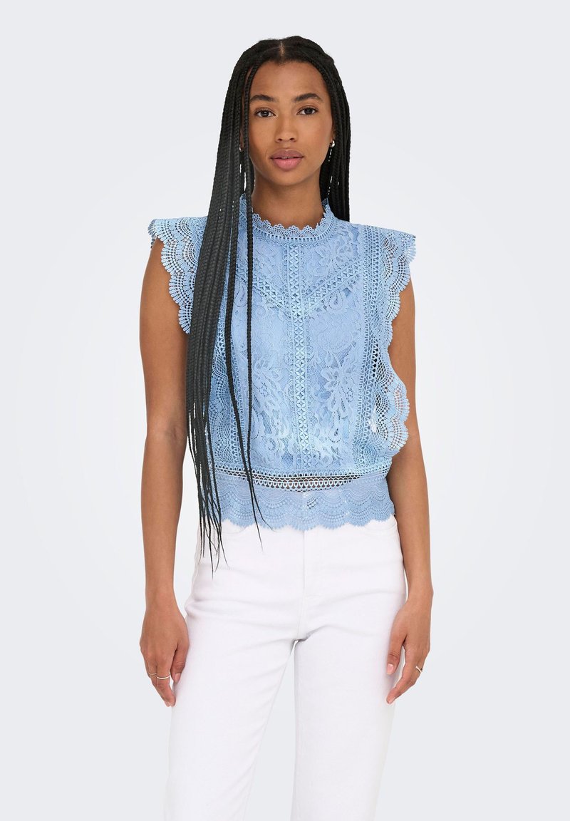 Mujer con trenzas largas, vistiendo una blusa sin mangas azul de encaje con bordes en forma de ondas y pantalones blancos, de pie contra un fondo claro.