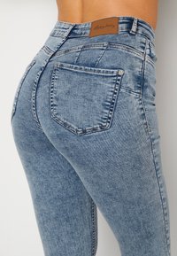 Jeans in denim blu con una silhouette aderente, dotati di due tasche posteriori e un'etichetta in pelle marrone in vita. Texture liscia e design casual.