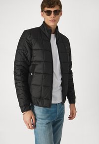 Jachetă neagră căptușită cu guler înalt, textură matlasată, buzunare laterale și închidere cu fermoar, purtată peste un tricou alb și blugi albaștri.