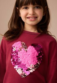 Felpa color bordeaux con un grande design a forma di cuore in paillettes nelle diverse sfumature di rosa e rosso. Texture liscia con dettagli luccicanti.