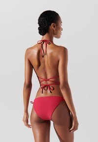 Bikini rosso con lacci al collo e schiena incrociata. Texture liscia; parte inferiore corta con lacci laterali. Design minimalista e colore vivace.
