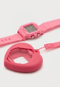 Ροζ ψηφιακό ρολόι χειρός με λουράκι από καουτσούκ τοποθετημένο δίπλα σε θήκη σιλικόνης σε σχήμα καρδιάς με υφασμάτινο λουράκι, σε λευκή επιφάνεια.