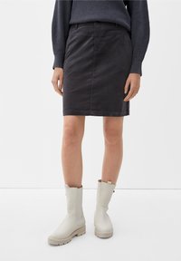 Jupe noire, de longueur genou, en tissu texturé avec une silhouette ajustée. Associée à des bottes en caoutchouc beige clair, mi-mollet, avec une semelle épaisse.