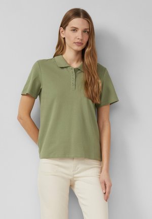 Kobieta stoi przy szarej ścianie, ubrana w jasnozieloną koszulkę polo z krótkim rękawem i kremowe spodnie, z długimi, prostymi włosami rozpuszczonymi.