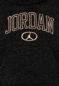 Černá mikina s kapucí. "JORDAN" v lesklých zlatých písmenech, s logem Jumpman v zlatém oválu. Měkká textura a přední kapsa.
