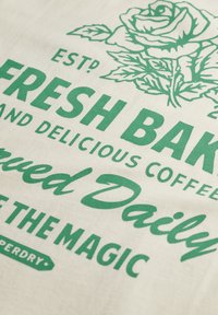 Bomuldsstof med grønt tekst, der siger "Friskbagt" og et rose-grafik. Har et vintage-design med en tekstureret, naturlig baggrund.