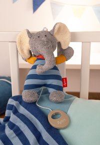 Elefante di peluche con grandi orecchie testurizzate, indossa un abito a righe blu. Vicino ci sono una morbida coperta blu e un attacco circolare.