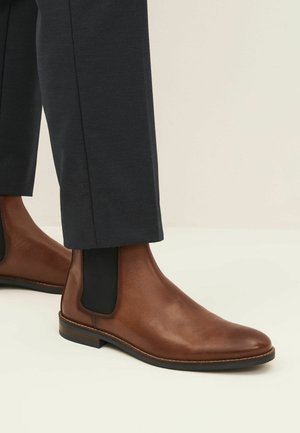 Botas Chelsea de cuero marrón con paneles laterales elásticos negros y puntera redondeada, combinadas con pantalones azul marino, con una textura suave y un tacón bajo.