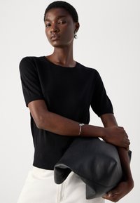 Black knit short-sleeve sweater with a ribbed hem, paired with a soft black leather clutch. Silver hoop earrings and a delicate bracelet are visible.