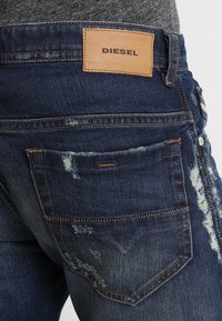 Jeans de mezclilla azul oscuro con un acabado desgastado, que presentan un parche de cuero etiquetado como "DIESEL" y bolsillos traseros estándar con detalles de costura.