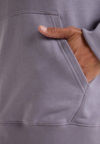 Felpa lavanda con tasca frontale e polsini a costine. La texture del tessuto sembra morbida e leggermente spazzolata, con dettagli delle cuciture ben visibili.