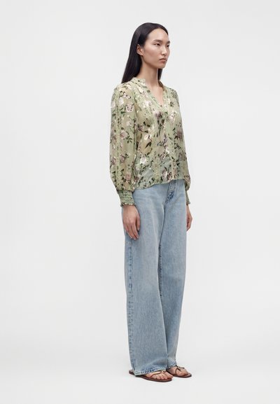Femme portant un chemisier vert transparent à motifs floraux, un jean large bleu clair et des sandales marron, debout devant un fond blanc uni.