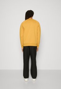 Persona con lunghi capelli intrecciati, che indossa un felpa senape, pantaloni cargo neri e sneakers bianche, in piedi di fronte a una parete grigia chiara uniforme.