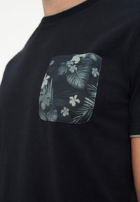 Κοντινό μιας μαύρης μπλούζας με σκούρο floral σχέδιο στην τσέπη στο στήθος που περιλαμβάνει φύλλα και λουλούδια.