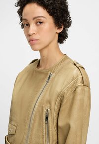 Veste en cuir beige avec une finition texturée, fermeture éclair asymétrique et épaulettes sur les épaules. Elle possède deux poches zippées.