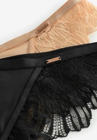 Melna un kailā krāsā apakšveļa no mežģīnēm un mīksta auduma, ar dekoratīvām mežģīņu malām un metāla zīmola birku.