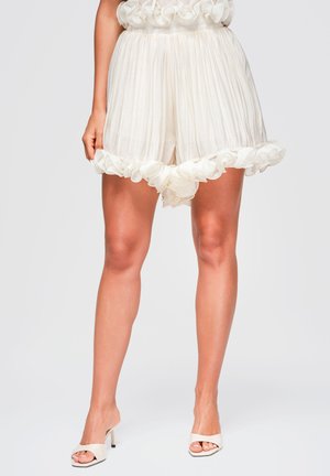 Femme portant un short blanc plissé avec un ourlet à volants et des sandales blanches à talons hauts ouverts, debout devant un fond uni.