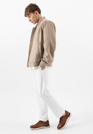 Mladý muž ve světle béžovém kabátu, bílých kalhotách a hnědých teniskách stojí s rukama v kapse a dívá se dolů.