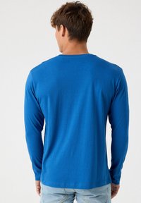 Camiseta de manga larga en tela azul vibrante, con un diseño de cuello redondo y textura suave. La vista trasera muestra simplicidad y líneas limpias.