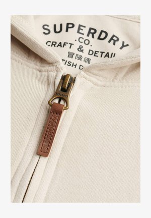 Close-up van een beige stoffen rits met een leren trekker met daarop "SUPERDRY" en zwart gedrukt tekst "SUPERDRY CO. CRAFT & DETAIL" op de binnenvoering.