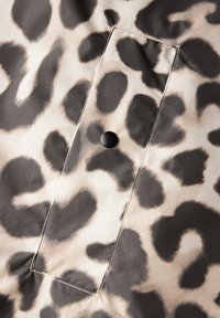 Tejido de estampado de leopardo con manchas negras sobre un fondo beige claro. Presenta un parche rectangular con un botón de presión negro. Textura suave.
