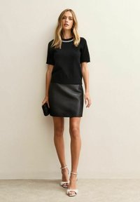 Zwarte trui met korte mouwen, gecombineerd met een zwart leren minirok met textuur en zilveren strappy hakken; houdt een kleine zwarte clutch vast.
