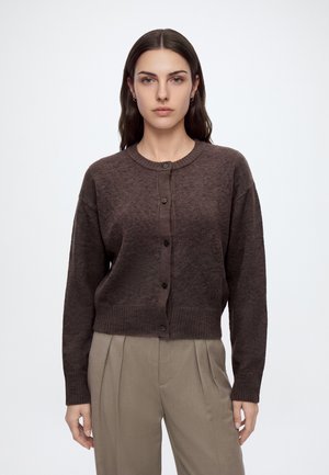 Vrouw met lang donker haar draagt een donkerbruine cardigan met knopen en beige geplooide broek, staand tegen een effen lichte achtergrond.