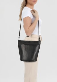 Czarna skórzana torba na ramię o minimalistycznym wzornictwie, płaskim kształcie i regulowanym pasku, wyeksponowana na neutralnym tle.