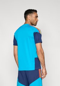 Athletisk t-shirt i klarblått och marinblått, med korta ärmar, rund halsringning och flerfärgade ränder längs axlarna.