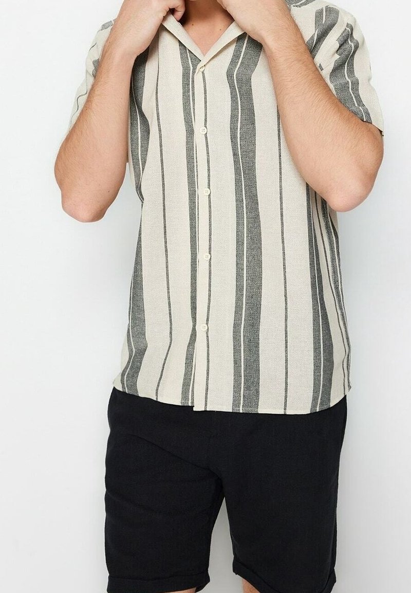 Man past steeds de kraag van een beige halfhande hemd met verticale zwarte strepen, draagt zwarte shorts tegen een witte achtergrond.