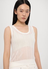 Balta bezpiedurkņu turtlenecka bluze ar adītu mežģīņu rakstu un ribinātu apakšmalu. Tam ir apaļa kakla izgriezums un nedaudz caurredzama tekstūra.