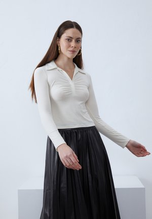 Langermet bluse i krem, med krage og rynket frontdetalj, sammen med et svart plisserte skjørt. Glatt tekstur.