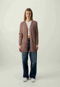 Cardigan de malha rosa claro com frente aberta, punhos canelados e dois bolsosfrontais, usado sobre uma t-shirt cropped branca e jeans azuis desfiados.