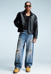 Man with braided hair and sunglasses wearing a black leather jacket, black shirt, ripped blue jeans, and tan boots standing against a light background.