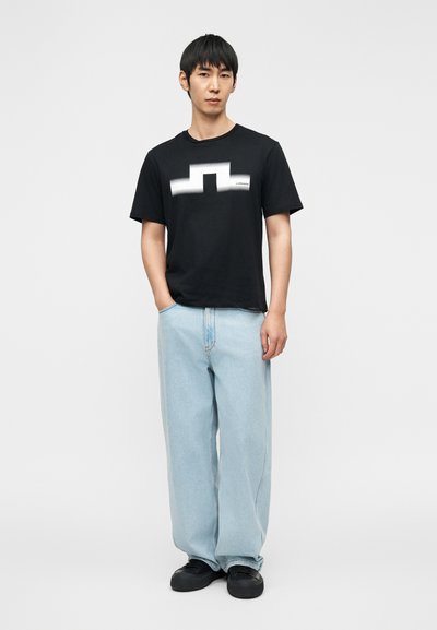 Νέος άνδρας που στέκεται με το ένα χέρι στην τσέπη, φοράει μαύρο γραφικό t-shirt, φαρδιά ανοιχτόχρωμα μπλε τζιν και μαύρα αθλητικά παπούτσια, μπροστά σε λευκό φόντο.