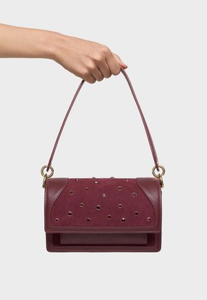 Main tenant un petit sac à main bordeaux avec un rabat clouté, des accents en cuir, des anneaux dorés, et une seule bandoulière sur un fond uni.