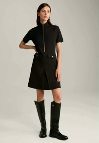 Zwarte jurk met een kraag met rits, korte mouwen en twee voorzakken. Gecombineerd met hoge zwarte leren laarzen met een zijsluiting.