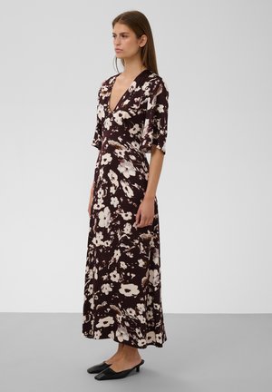 Femme debout de profil portant une longue robe fleurie sombre avec un profond décolleté en V et des manches jusqu'aux coudes, chaussures noires à petit talon, fond neutre.