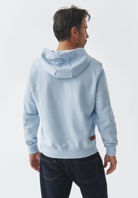 Sudadera azul claro con capucha, puños y dobladillo acanalados, detalle de parche de cuero en la parte posterior y textura suave. Llevada con jeans oscuros.