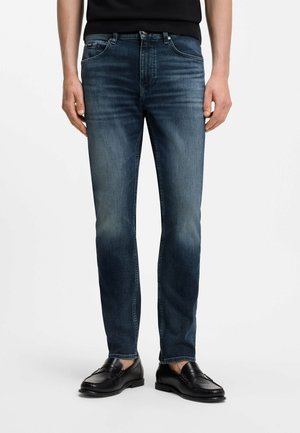 Mężczyzna noszący ciemnoniebieskie jeansy o kroju slim fit i czarne skórzane mokasyny, stojący na jednolitym jasnym tle.