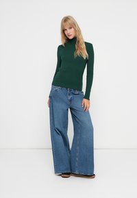 Zaļš garo piedurkņu turtlenecks, apvienots ar platām zilo džinsu biksēm. Mīksts audums, brīvs piegriezums un izteikta šuvju detaļas uz biksēm.