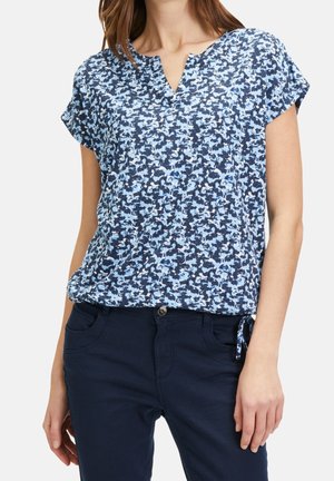 Femme portant un chemisier à manches courtes bleu et blanc à motif, avec un col cranté, associé à un pantalon bleu foncé.