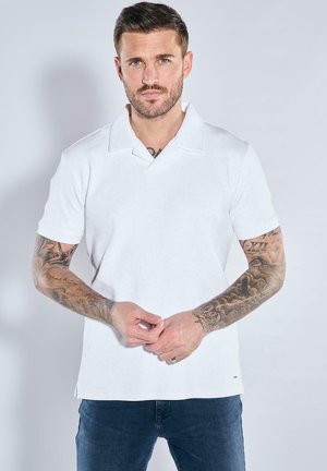 Weißes Polohemd mit kurzen Ärmeln und gerippter Textur, flachem Kragen und seitlichen Schlitzen, kombiniert mit dunkelblauen Jeans und tätowierten Armen.