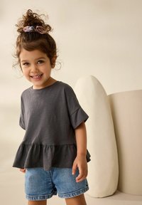 Haut gris à manches courtes avec un ourlet en peplum, associé à un short en denim clair. L'arrière-plan présente des formes douces et de couleurs neutres.