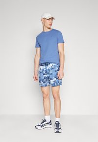 Camiseta de algodón azul, pantalones cortos azules con un diseño tropical y zapatillas blancas. El modelo lleva una gorra blanca. Look sencillo y casual.