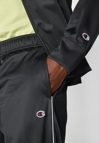 Černá sportovní bunda se zipem v přední části, v kombinaci s odpovídajícími kalhotami. Obsahuje malé vyšívané logo a světlé akcenty.