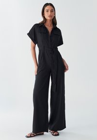 Juodas kombinezonas su trumpomis rankovėmis, priekine sagų eilute, dviem krūtinės kišenėmis ir surišamu juosmeniu. Pagamintas iš lengvos medžiagos su plačiomis kelnėmis.