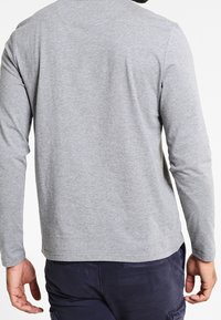 Man i ljusgrå långärmad skjorta och mörkblå byxor, vänd bort med händerna avslappnat längs sidorna.